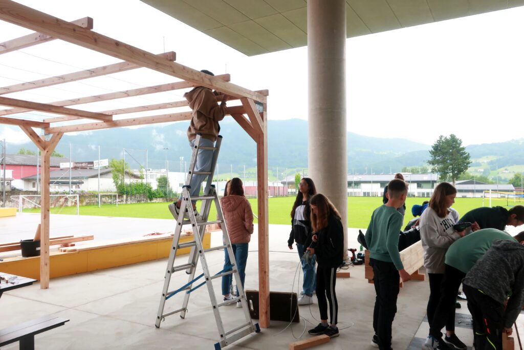 Im Bild sind Schüler*innen zu sehen, die verschiedene Montagearbeiten an einer Holzpergola vornehmen. Eine Person steht auf einer Leiter und befestigt ein Rankseil an der Konstruktion.