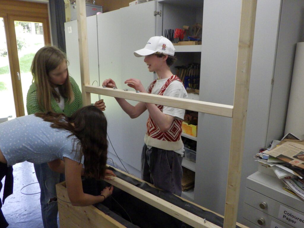 Im Bild sind Schüler*innen zu sehen, die Stahlseile als Rankhilfe an einem Pflanztrog aus Holz montieren.