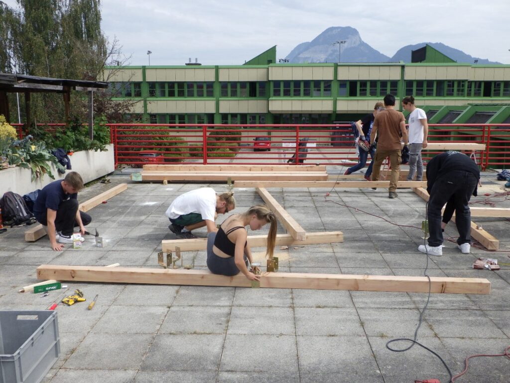 Im Bild sind Schüler*innen zu sehen, die unter Anleitung verschiedene Arbeiten am Aufbau einer Holzkonstruktion vornehmen.