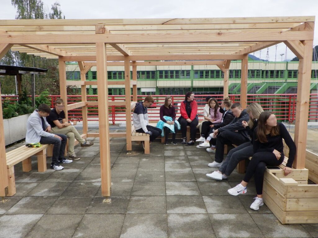 Auf dem Bild sind Jugendliche einer Schulklasse zu sehen, die unter einer Pergola aus Holz sitzen.
