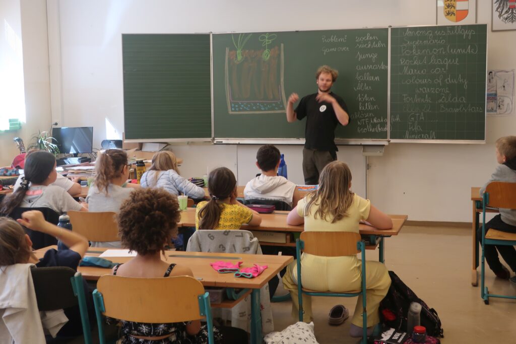 Im Bild sind sitzende Schüler*innen im Klassenzimmer und ein Vortragender an der Tafel zu sehen.