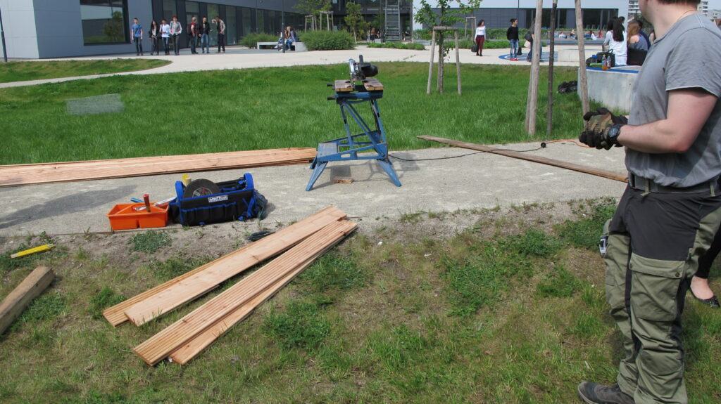 Im Bild sind Holzbretter und Bearbeitungsgeräte in einem Garten zu sehen.