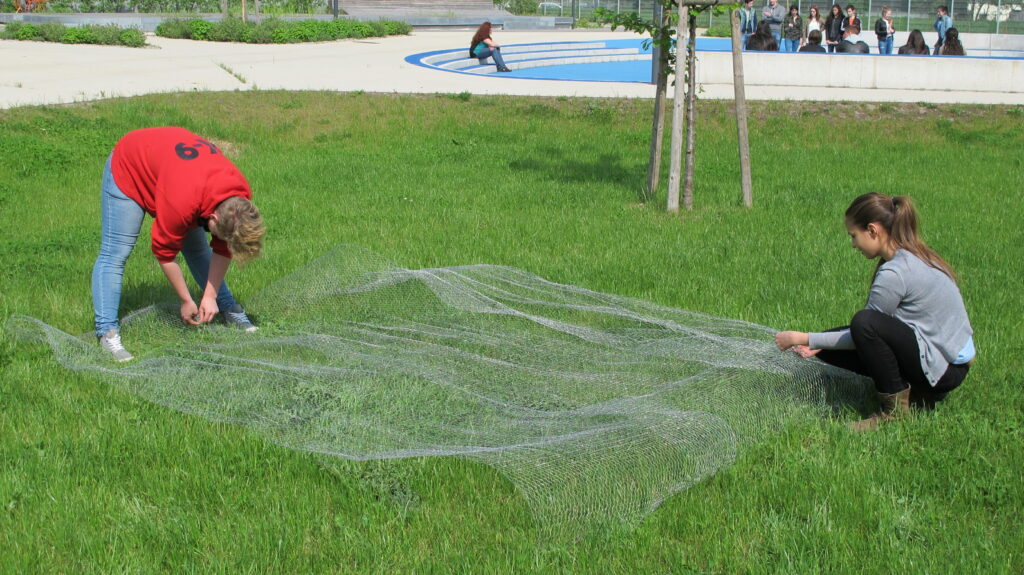 Im Bild sin zwei Schüler*innen zu sehen, die ein Drahtnetz auf der Wiese entfalten.